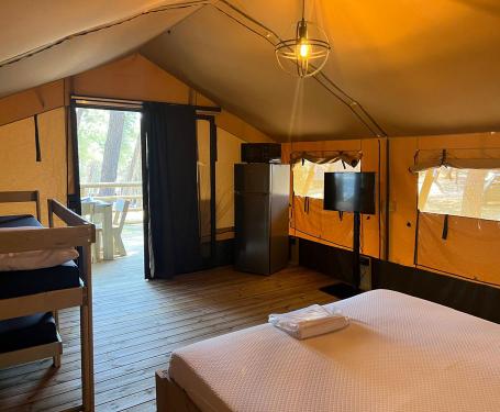 Glamping-Zelt mit Bett, Kühlschrank, Fernseher und Veranda inmitten der Natur.